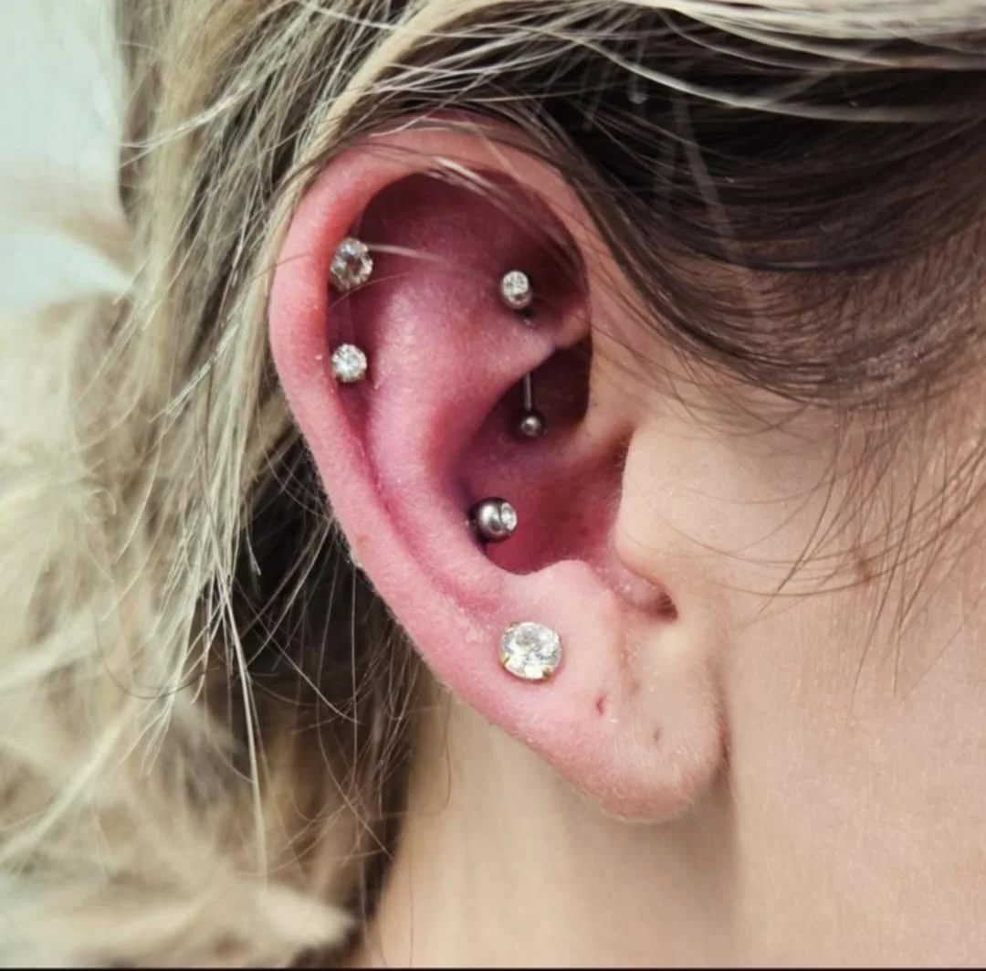 Close-up of an ear with multiple piercings, featuring several diamond studs and a circular barbell. The ear has a mix of lobe and cartilage piercings.