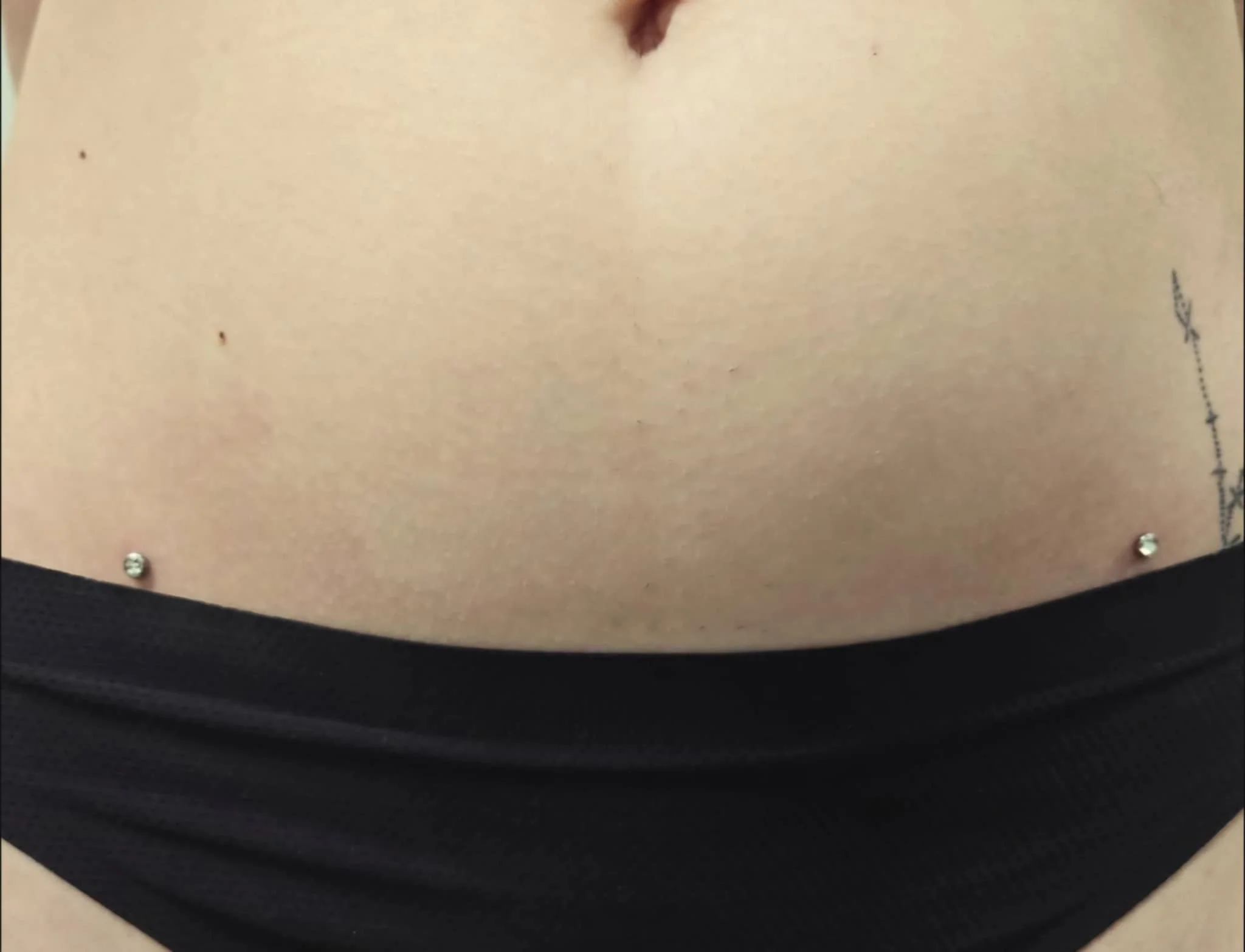 Close-up of a person's lower abdomen with hip piercings and a black garment.