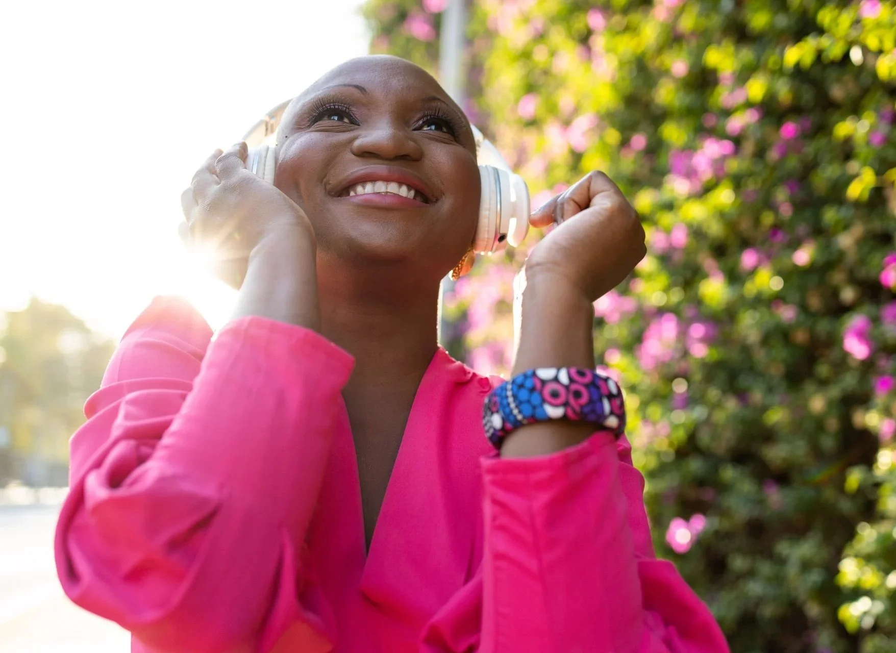A joyful woman with a bright smile, wearing a pink jacket, enjoying music with white headphones outdoors during daylight, with pink flowers and greenery in the background.