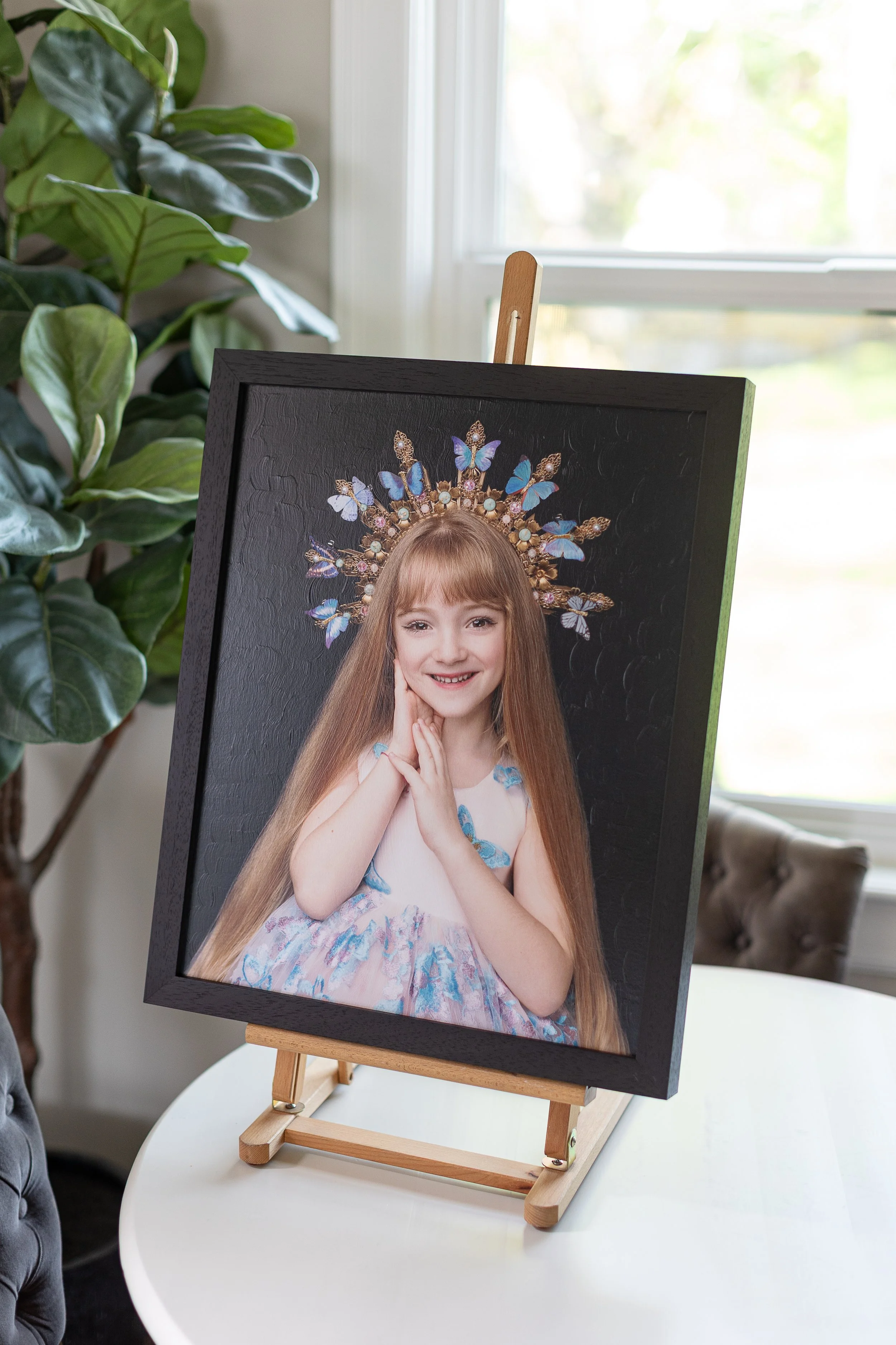 Framed portrait of a young girl with long red hair, wearing a pastel dress with blue butterfly accents and a decorative butterfly halo, on a small easel in a room with large window and green plant.