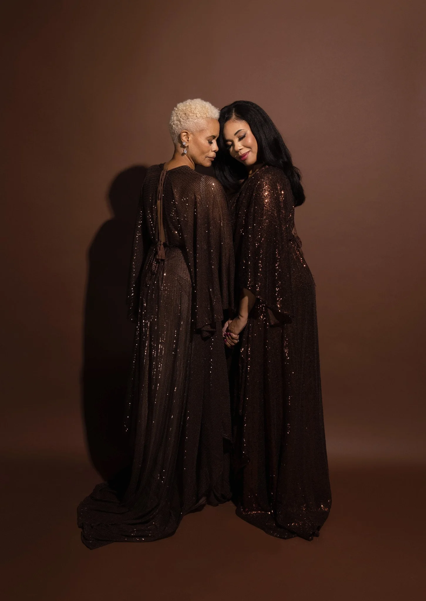 Three women of different ages, dressed in elegant, sequined dresses, smiling and embracing each other against a neutral background.