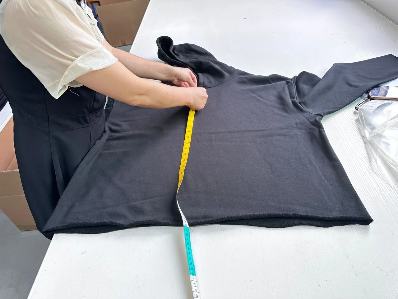 Person measuring a black hoodie on a white table with a tape measure.