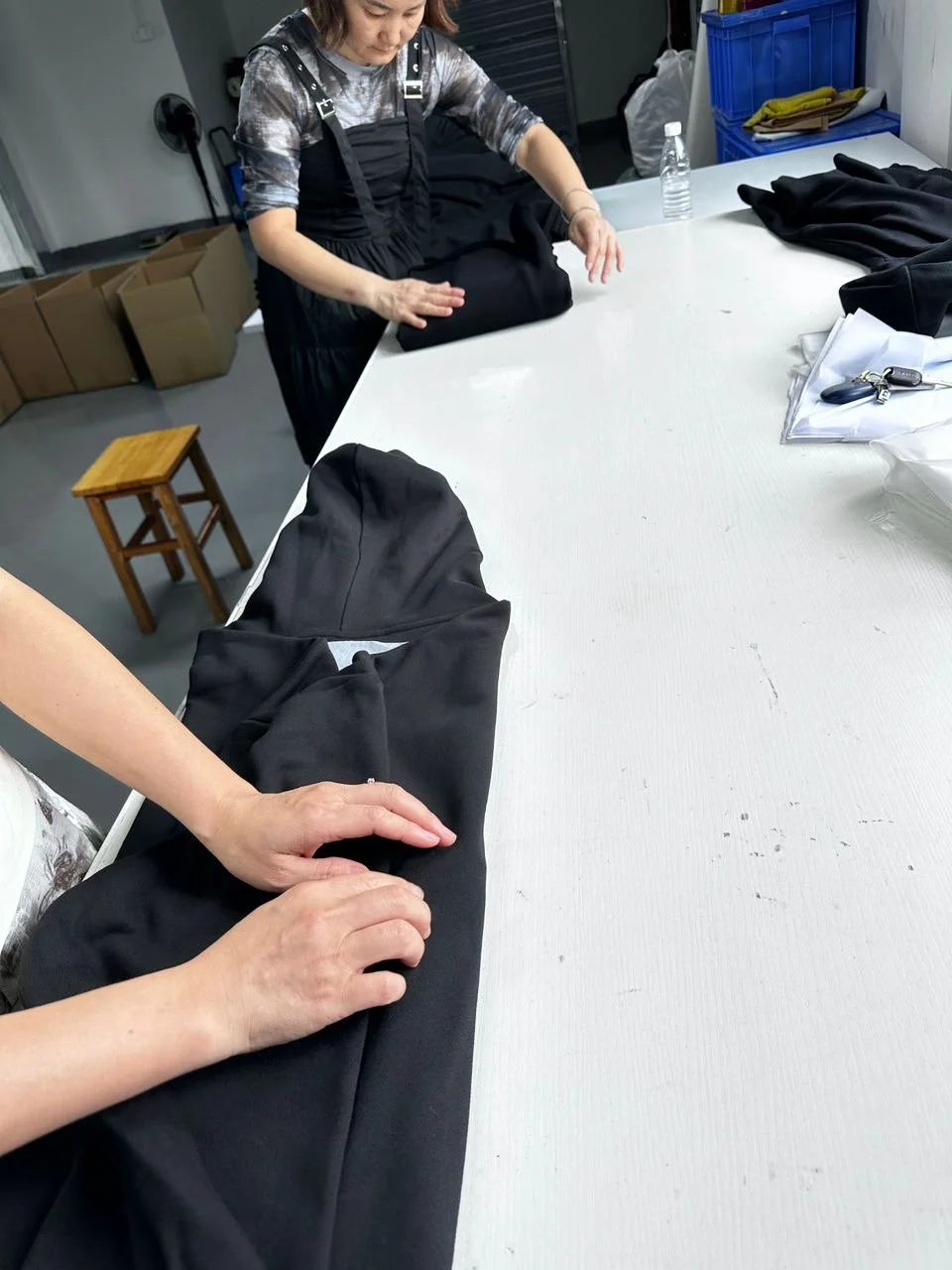 Two women are folding black clothing on a white table in a room with storage boxes and a water bottle.