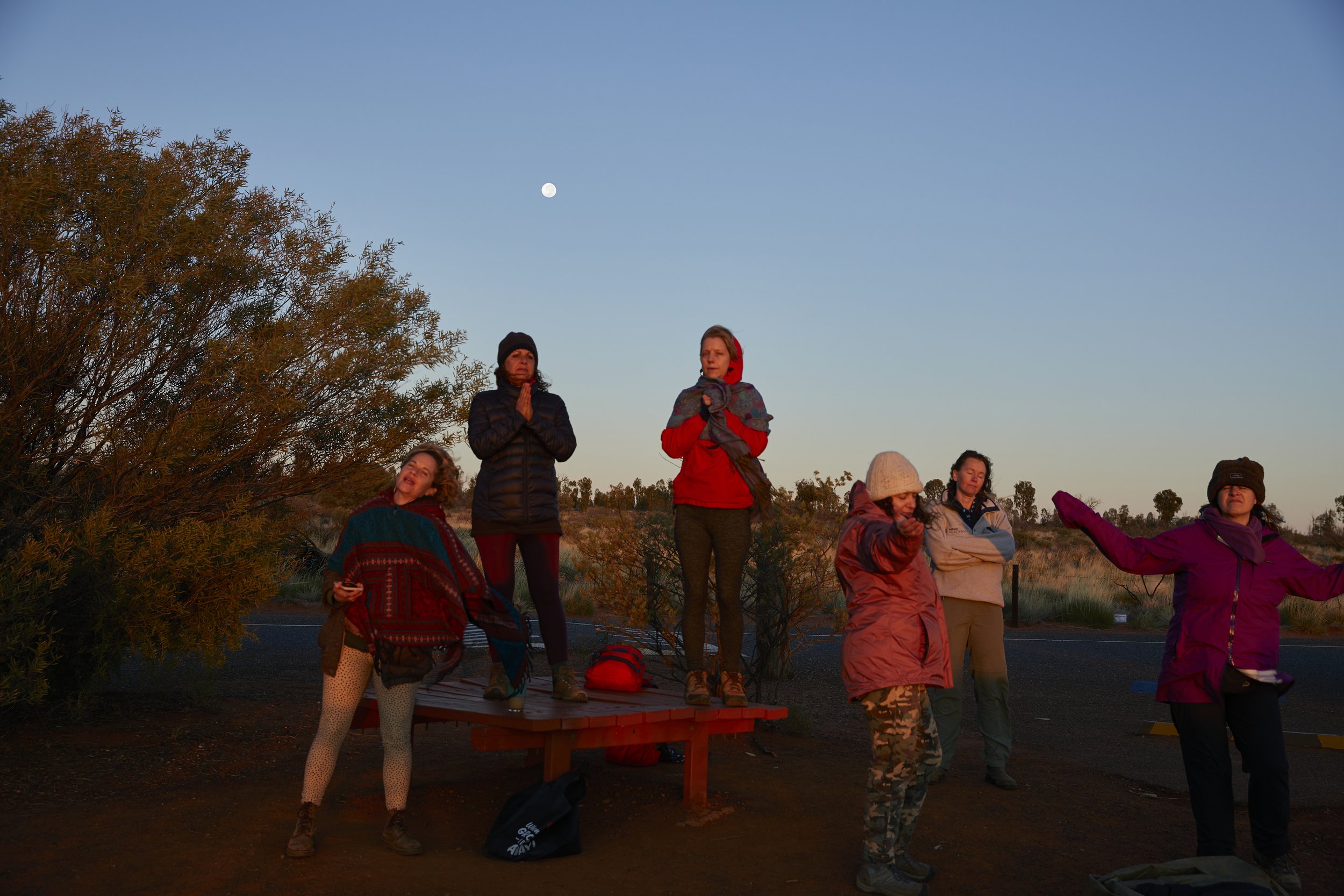 A group of people standing and sitting outdoors during dusk, some with hands in prayer position, with a clear sky and full moon in the background.