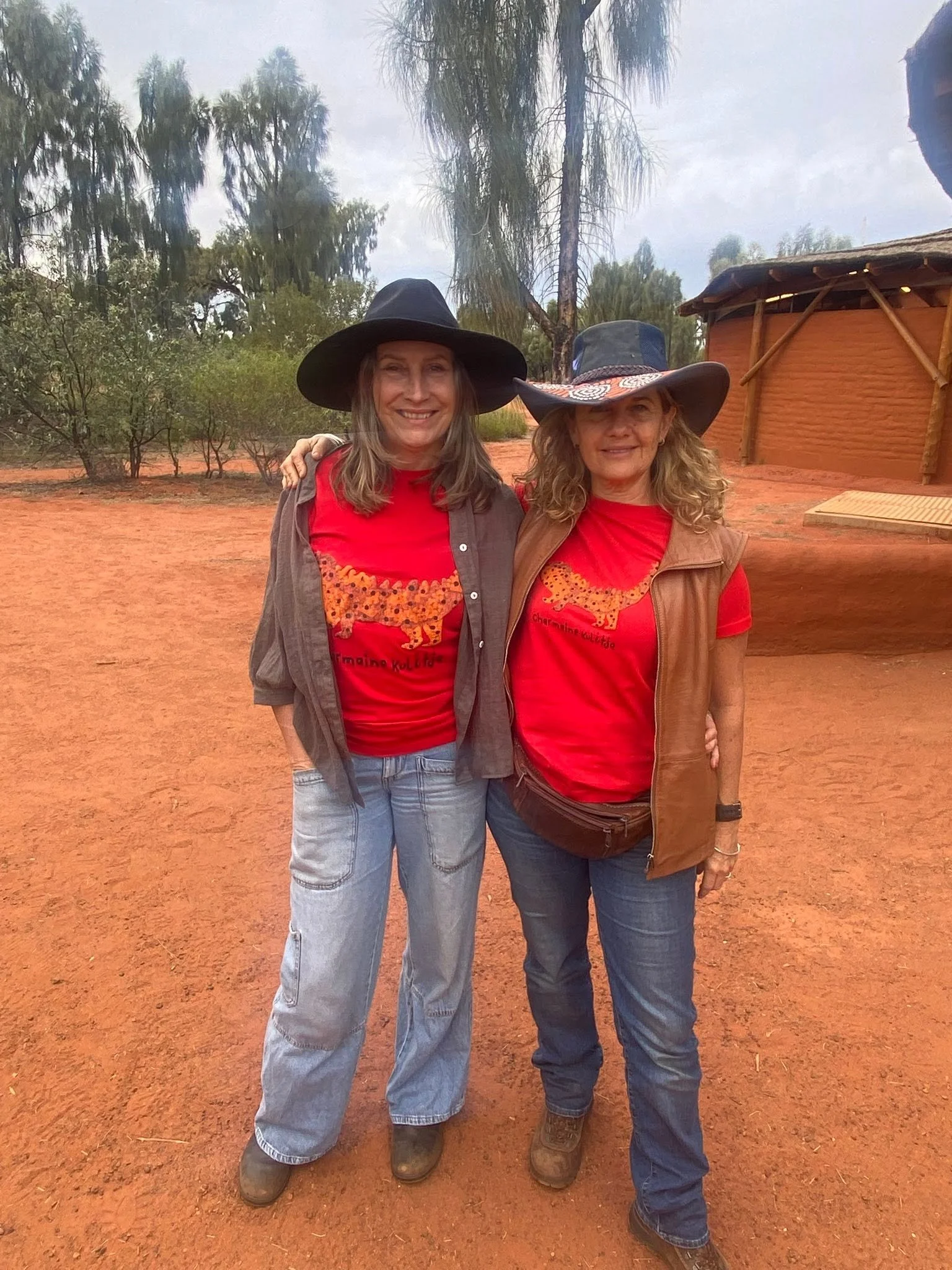Iris Ray Nunn and Serana Hunt-Hughes standing outdoors on central australia red dirt ground, smiling, wearing wide-brimmed hats and red shirts with a yellow lion design.  There are trees and a traditional round hut with a thatched roof in background.