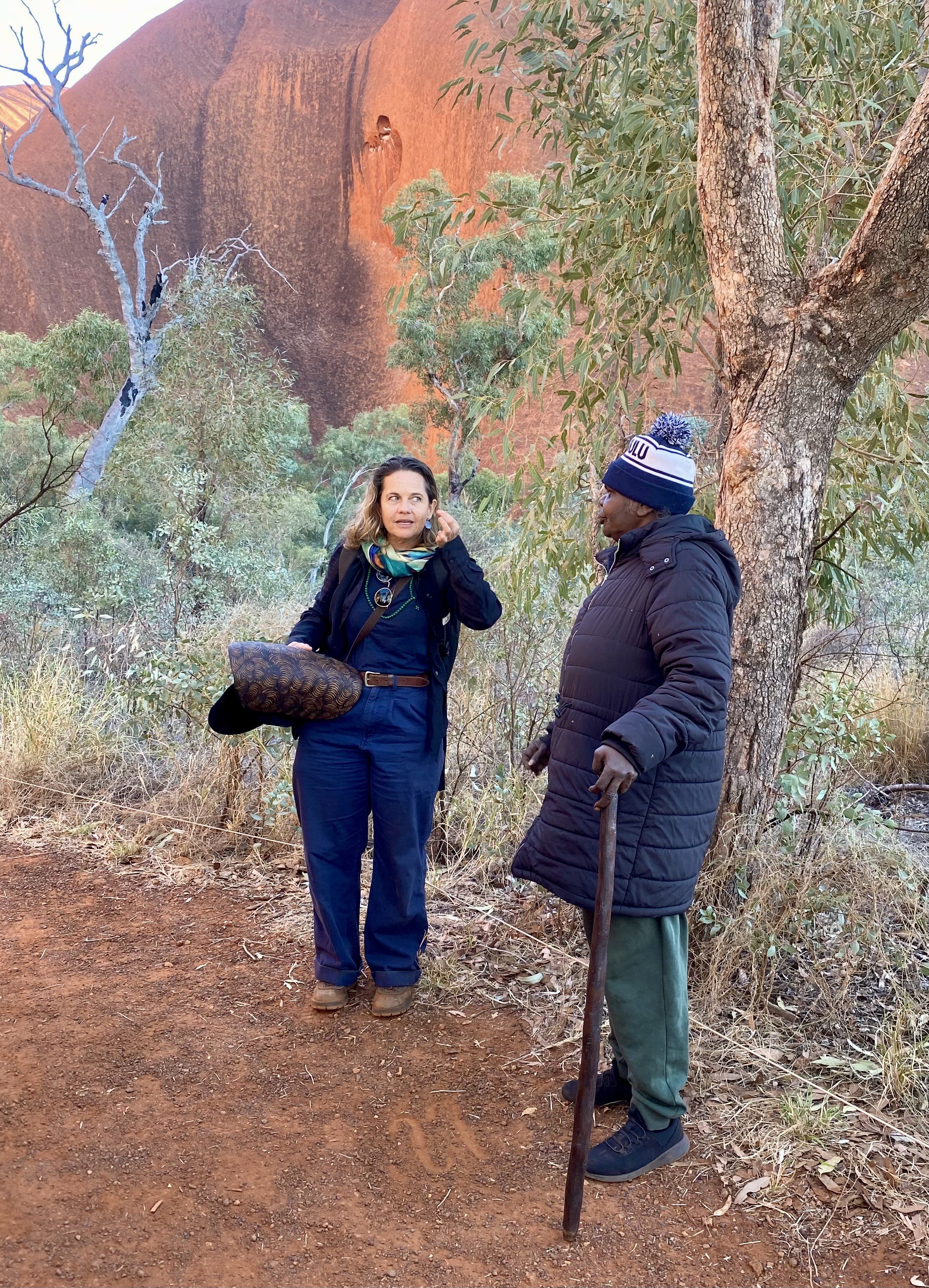 Two women talking outdoors near a tree, with a red rock formation in the background. One woman with blonde hair, wearing a black jacket, blue scarf, and carrying a patterned bag, is looking at the other woman. The other woman has dark skin, gray hair