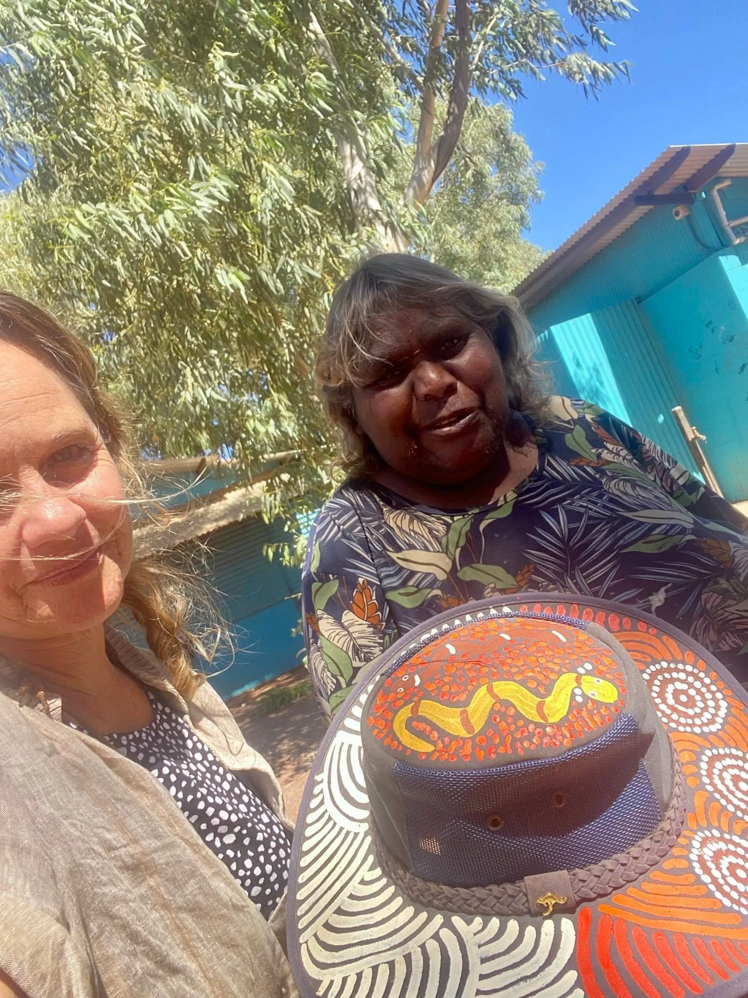 Iris Ray Nunn and Aṉangu friend taking a selfie in central australia. one with blonde hair and the other with darker hair, sitting at a table with a colorful indigenous art painted har, under a tree with green leaves and a bright blue sky.