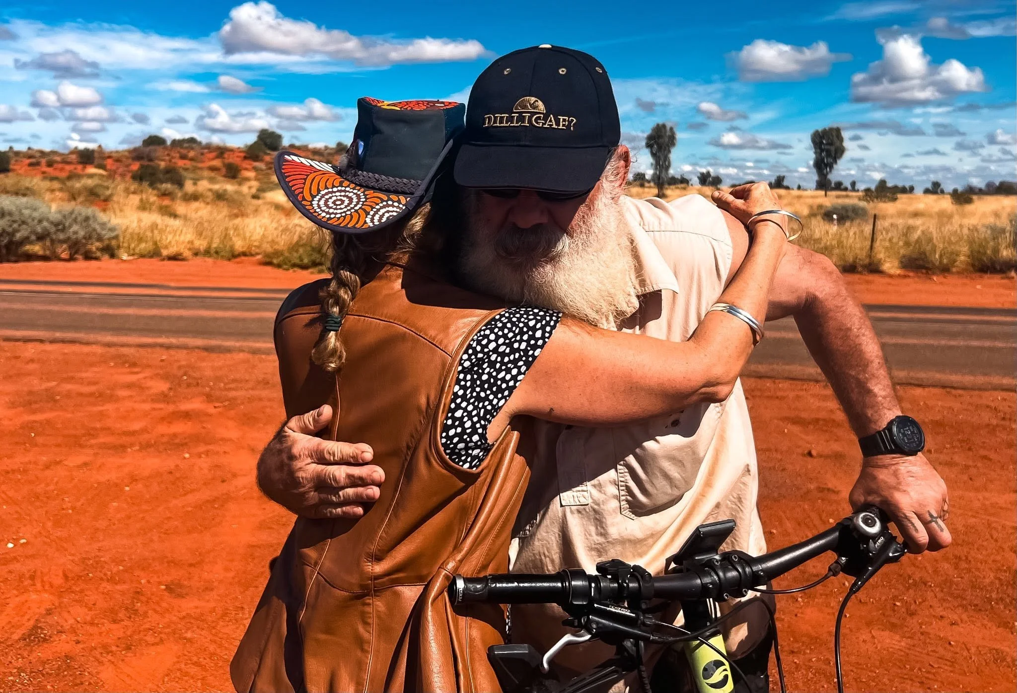 An Aṉangu man with a white beard and long hair, wearing a black cap, is stopped from riding a bicycle and hugging nature guide Iris. Central Australia desert landscape with red sand, sparse bushes, and a blue sky with white clouds. 