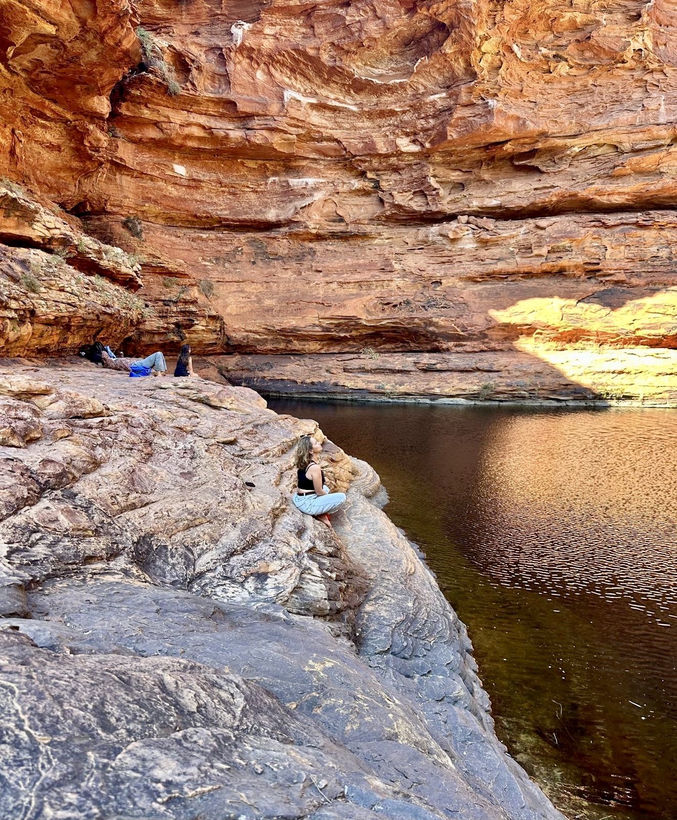 Group of people sitting and relaxing on rocks near a body of water inside a large central australia canyon or rock formation with reddish-orange walls.