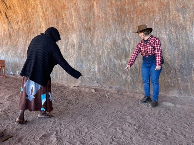 Two women are in a dirt-lined room with a bare, textured wall. One woman on the left, dressed in a black sweater and a colorful skirt, is holding a whip or riding crop. The other woman on the right, wearing a cowboy hat, glasses, plaid shirt, and jeans, is standing with her arms at her sides, looking at the woman with the whip.