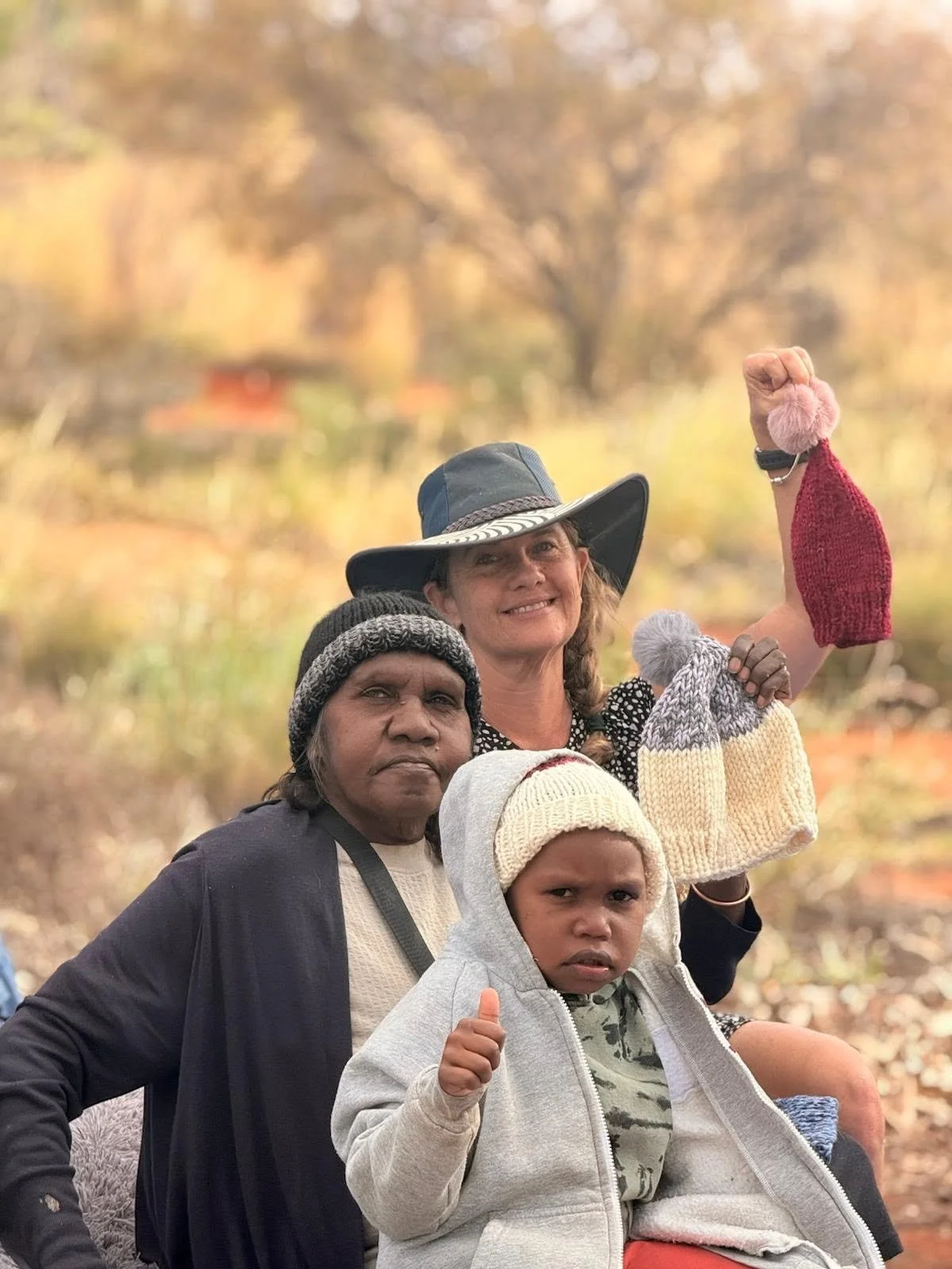 Iris Ray Nunn and Aṉangu friends. Group of four people outdoors in fall, three women and a young boy, wearing casual fall clothing and hats, with background of trees with autumn leaves.