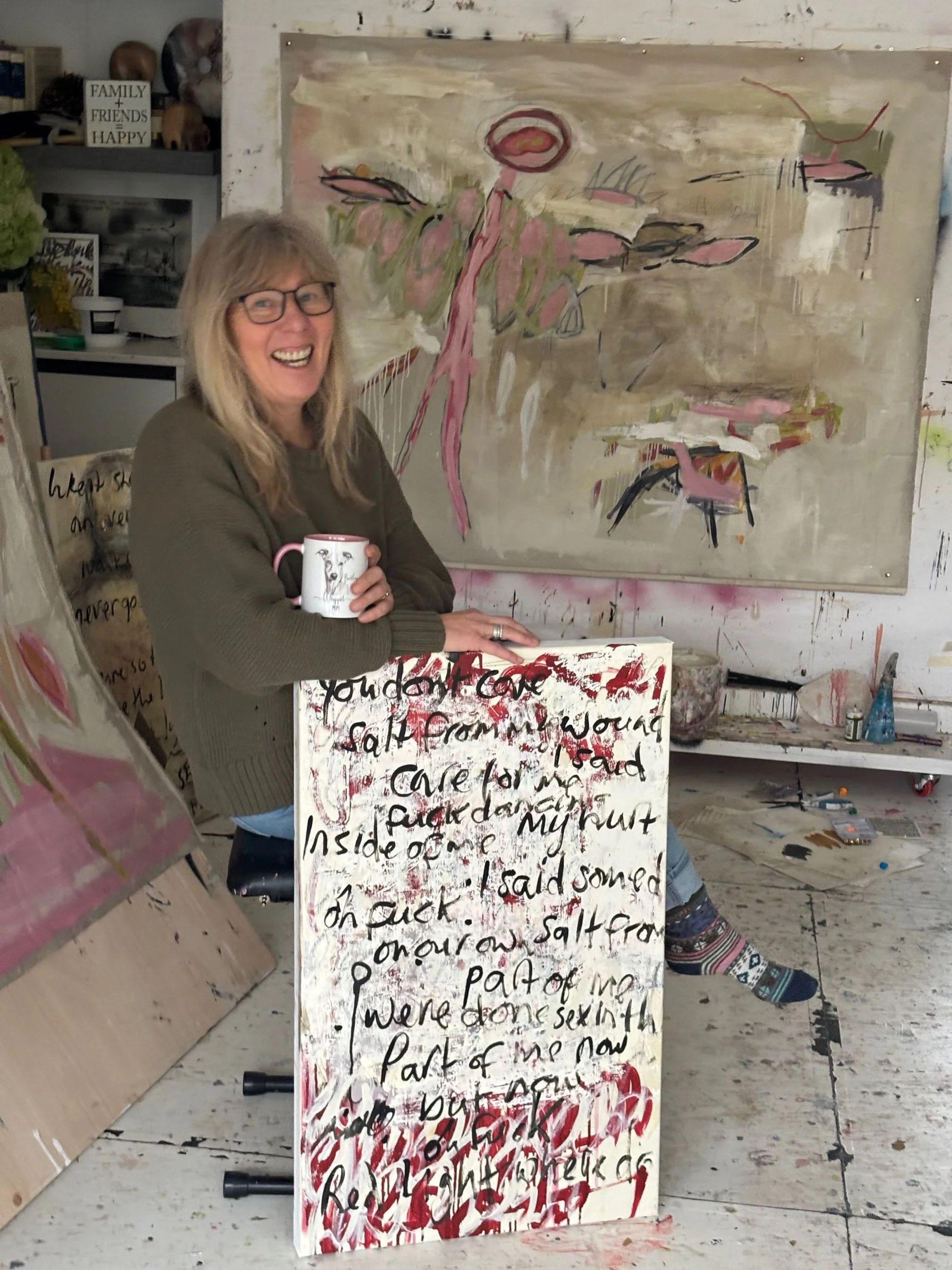 A woman with glasses and long blonde hair smiling in an art studio, holding a decorated mug. Behind her is a large abstract painting with pink and beige hues, and she is sitting next to a canvas with handwritten text and red paint, leaning on an easel. The studio has shelves with art supplies and a small sign that reads 'Family + Friends = Happy'.