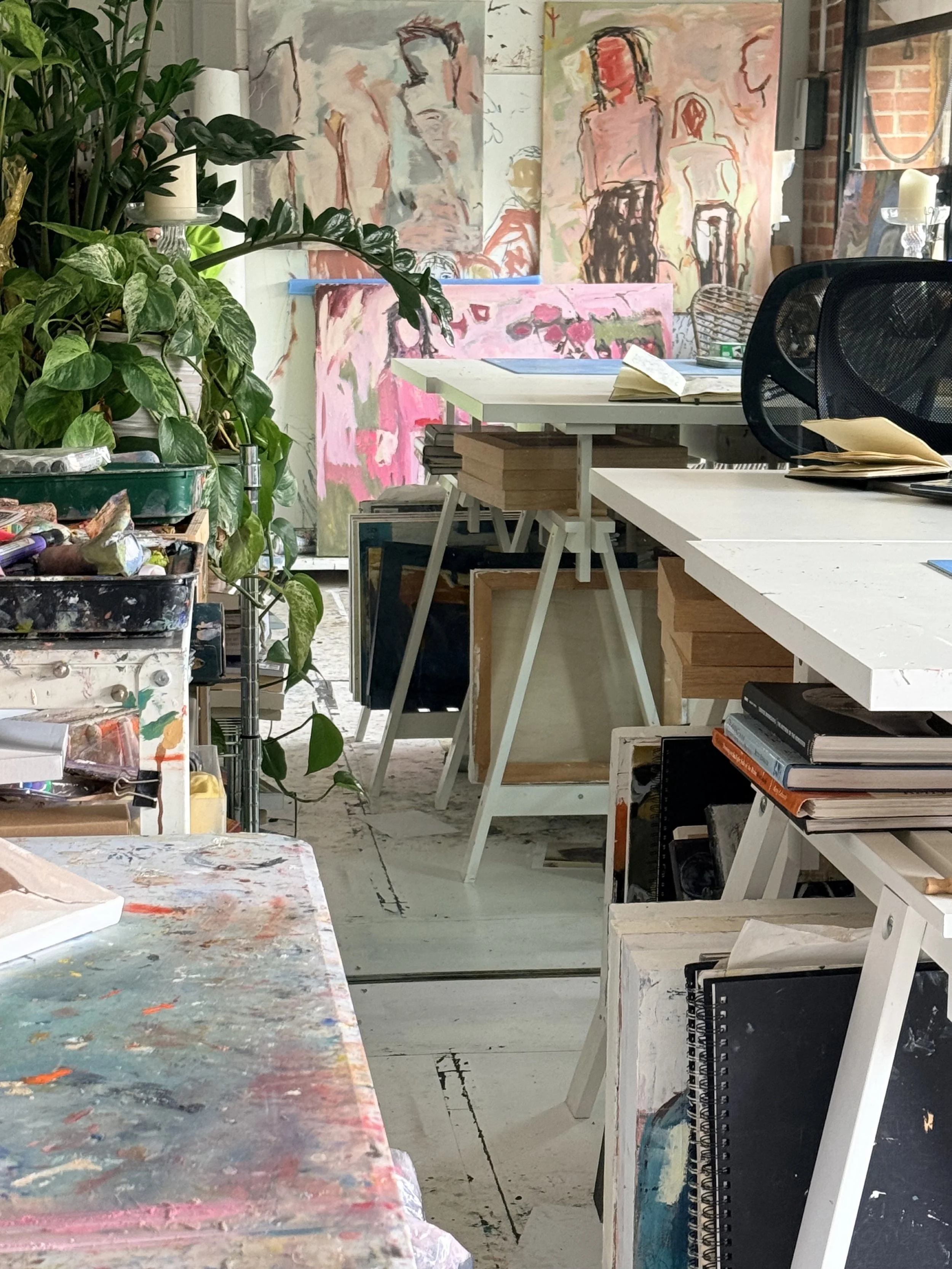 An artist's studio with colorful paintings on the wall, art supplies on a table, books, and large leafy green plants, with a window on the right side.