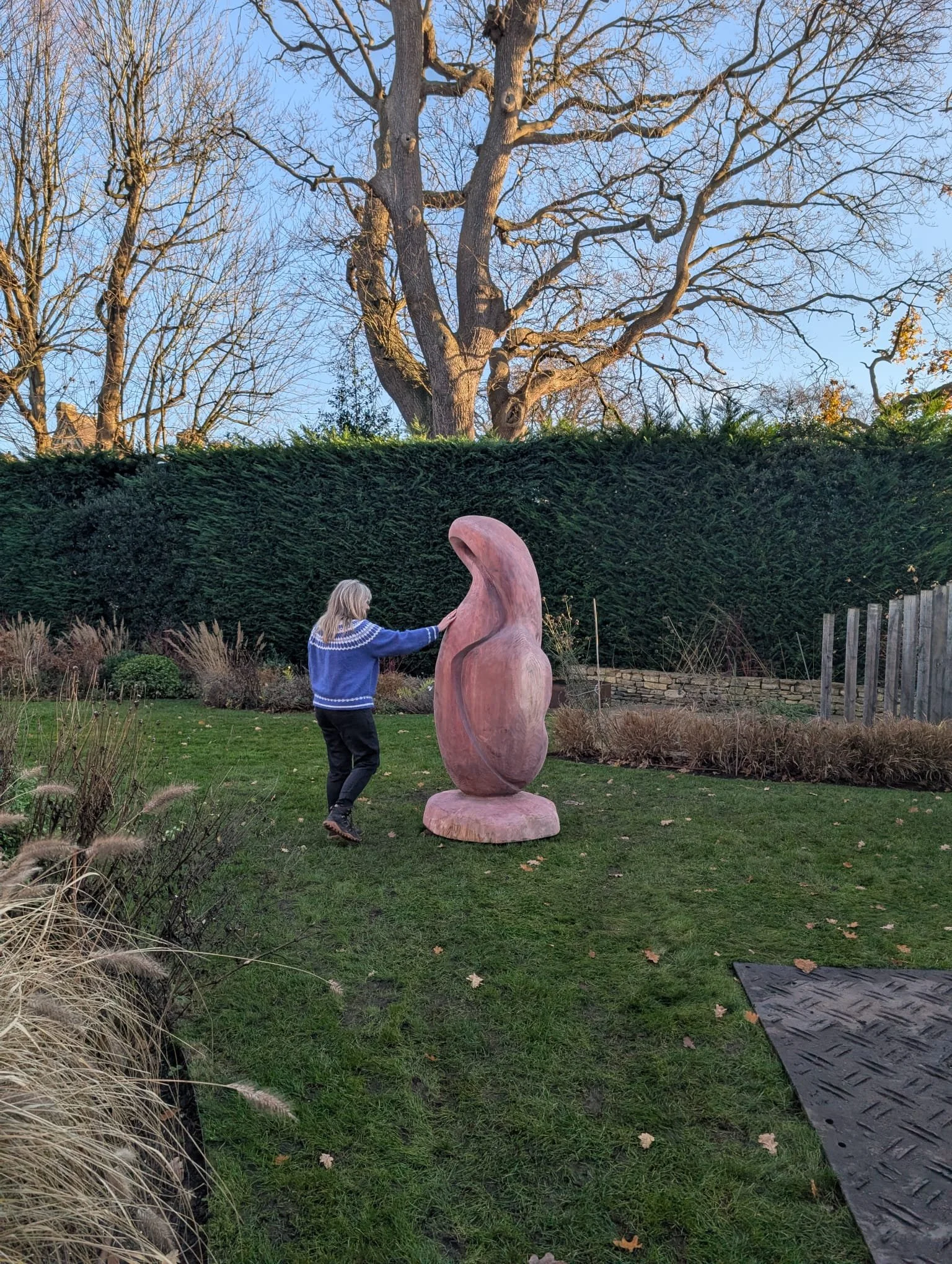 A woman is touching a large abstract wooden sculpture in a garden with grass, plants, and a tall hedge, under a clear blue sky and leafless trees.