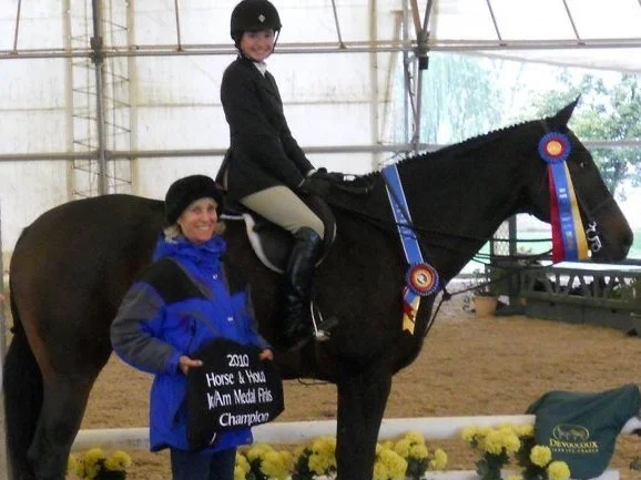 A young girl in equestrian riding attire sitting on a black horse, with a woman in a blue jacket holding a plaque that reads '2010 Horse & Hound AM Medal Finals Champion.' The horse has ribbons attached to its bridle, inside an indoor riding arena wi