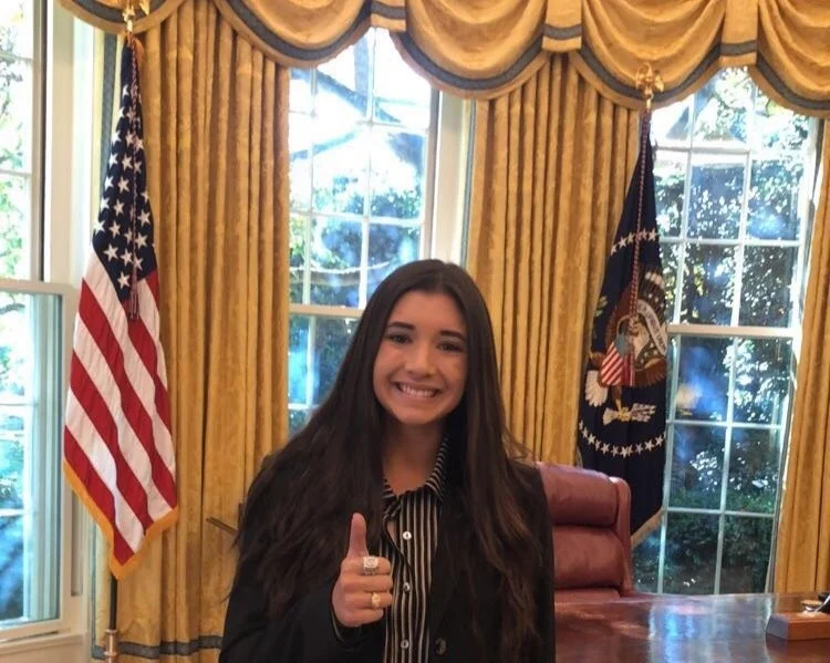 Young woman with long dark hair smiling and giving a thumbs up in the Oval Office, with American flags and gold curtains in the background.