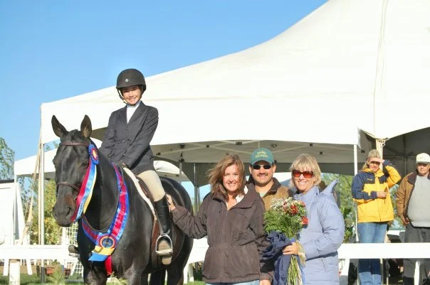 A young girl in riding gear on a horse with a ribbon, four adults smiling, one holding a bouquet of flowers, at an outdoor event under a large white tent.
