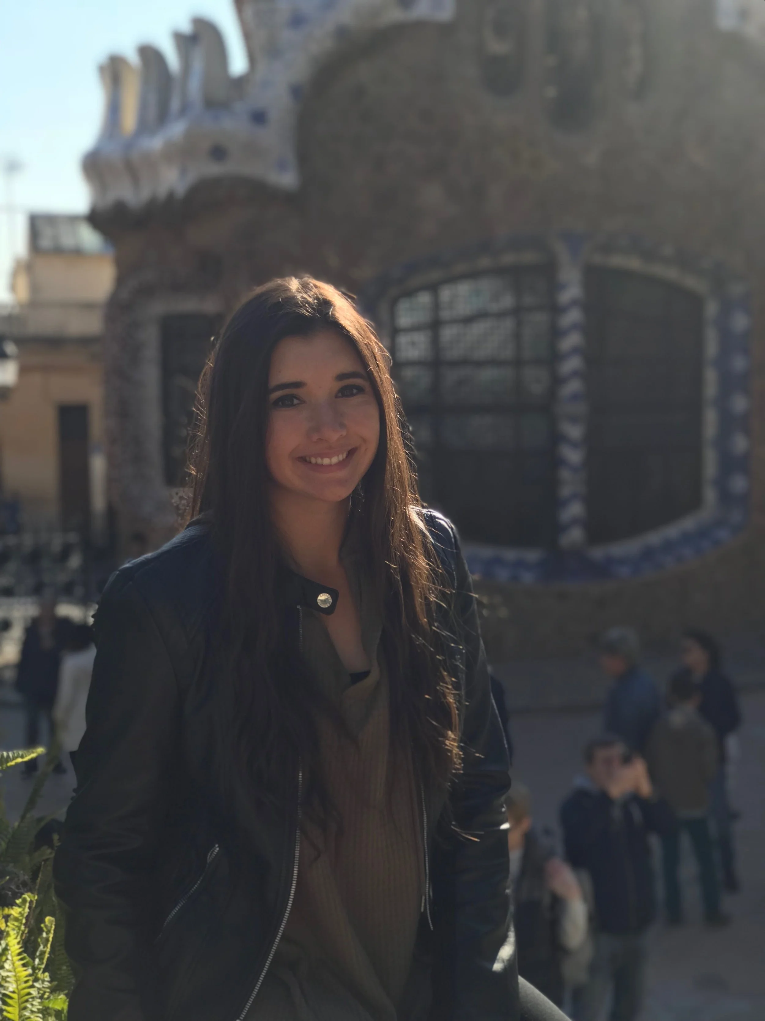 Smiling woman with long brown hair, wearing a black leather jacket, outdoors in front of a building with mosaic details.