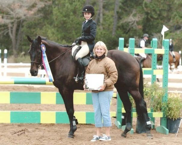 Young girl in riding gear on a horse with ribbon, standing next to a smiling woman holding a plaque at an equestrian competition.