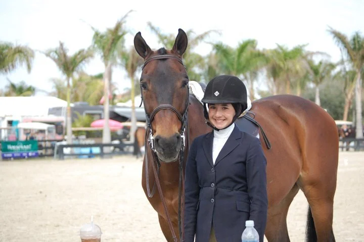 Young girl in riding gear and helmet standing next to a brown horse with a bridle at an outdoor equestrian event with palm trees and tents in the background.