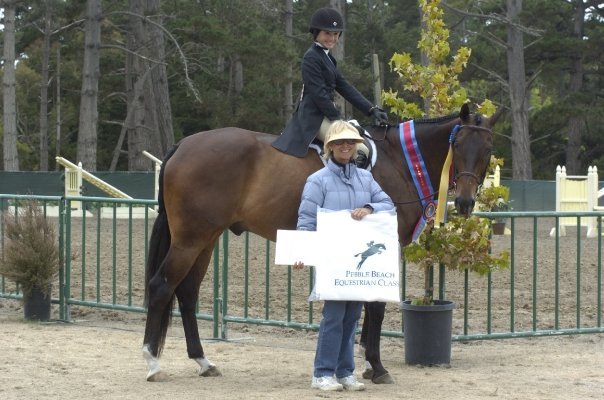 A young girl in a riding helmet sits on a brown horse with a blue ribbon, while a woman in sunglasses holds a white sign with a horse logo and stands next to them. The scene is outdoors with a fenced riding arena and trees in the background.
