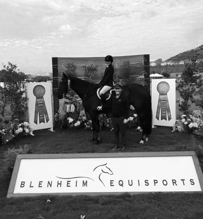Person riding a horse at an equestrian event with awards, flowers, and a banner for Blenheim Equisports in the background.
