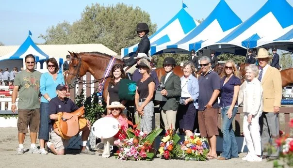 Group of people at an equestrian event with a horse, blue and white tents, and floral arrangements.