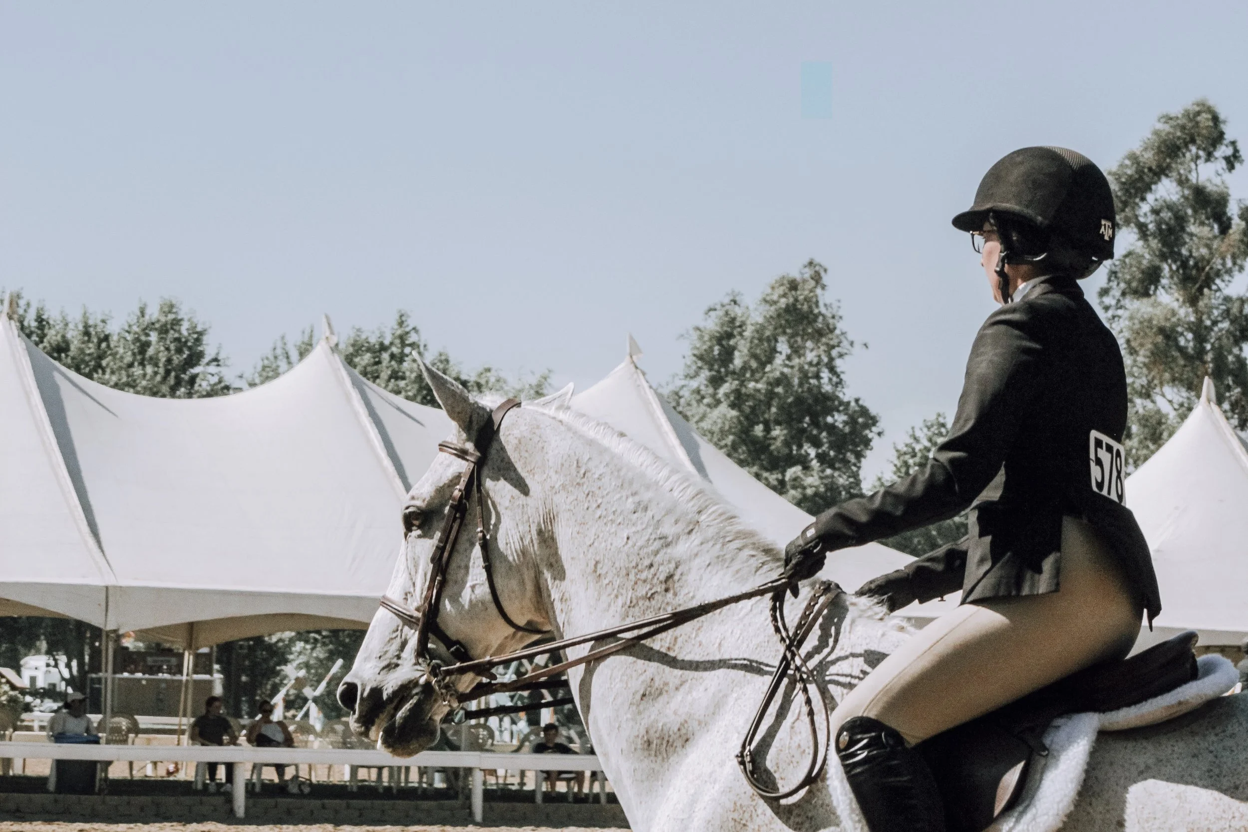 A rider in equestrian competition, wearing a black helmet and jacket, riding a white horse with a speckled coat, under large white tents with trees in the background.