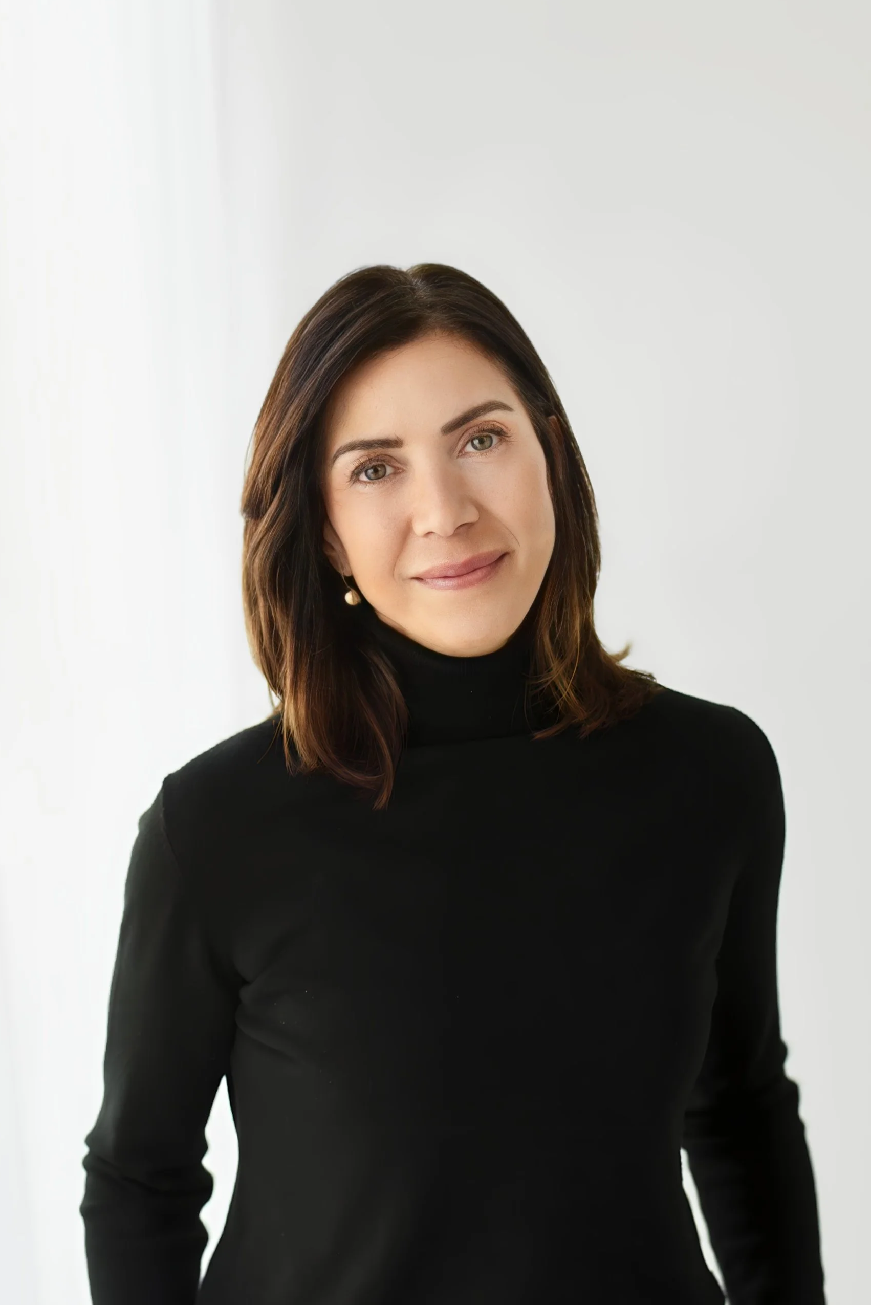 A woman with shoulder-length dark brown hair, wearing a black turtleneck sweater, smiling softly, standing against a plain light-colored background.