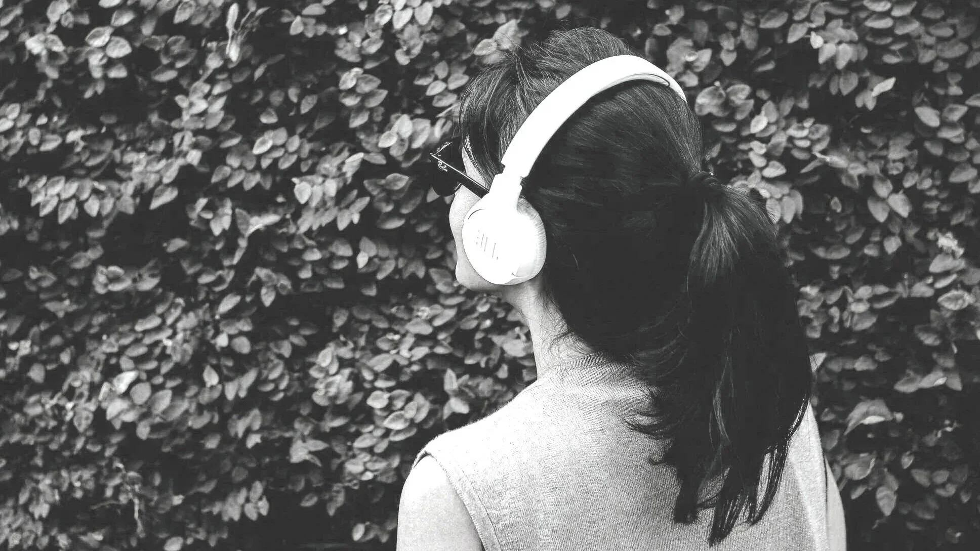 A woman with long, dark hair wearing large headphones, viewed from behind, standing in front of a dense hedge of leaves.