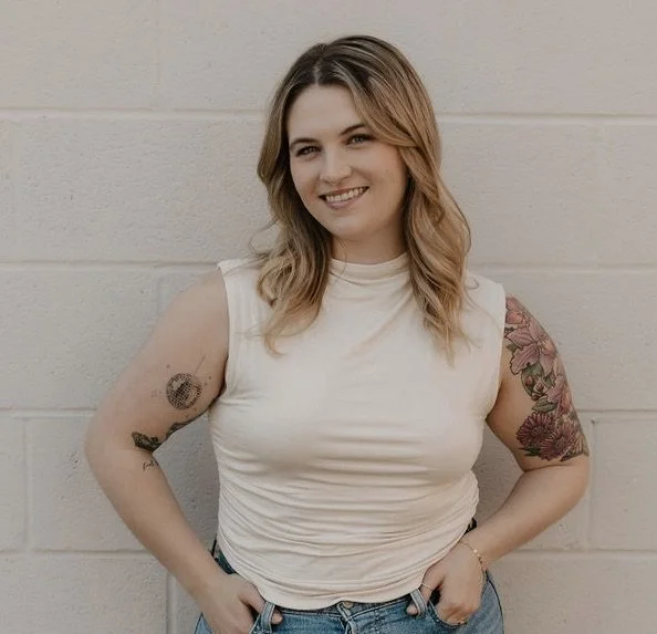 A smiling woman with shoulder-length wavy blonde hair, wearing a sleeveless white top and blue jeans, standing against a light-colored brick wall.