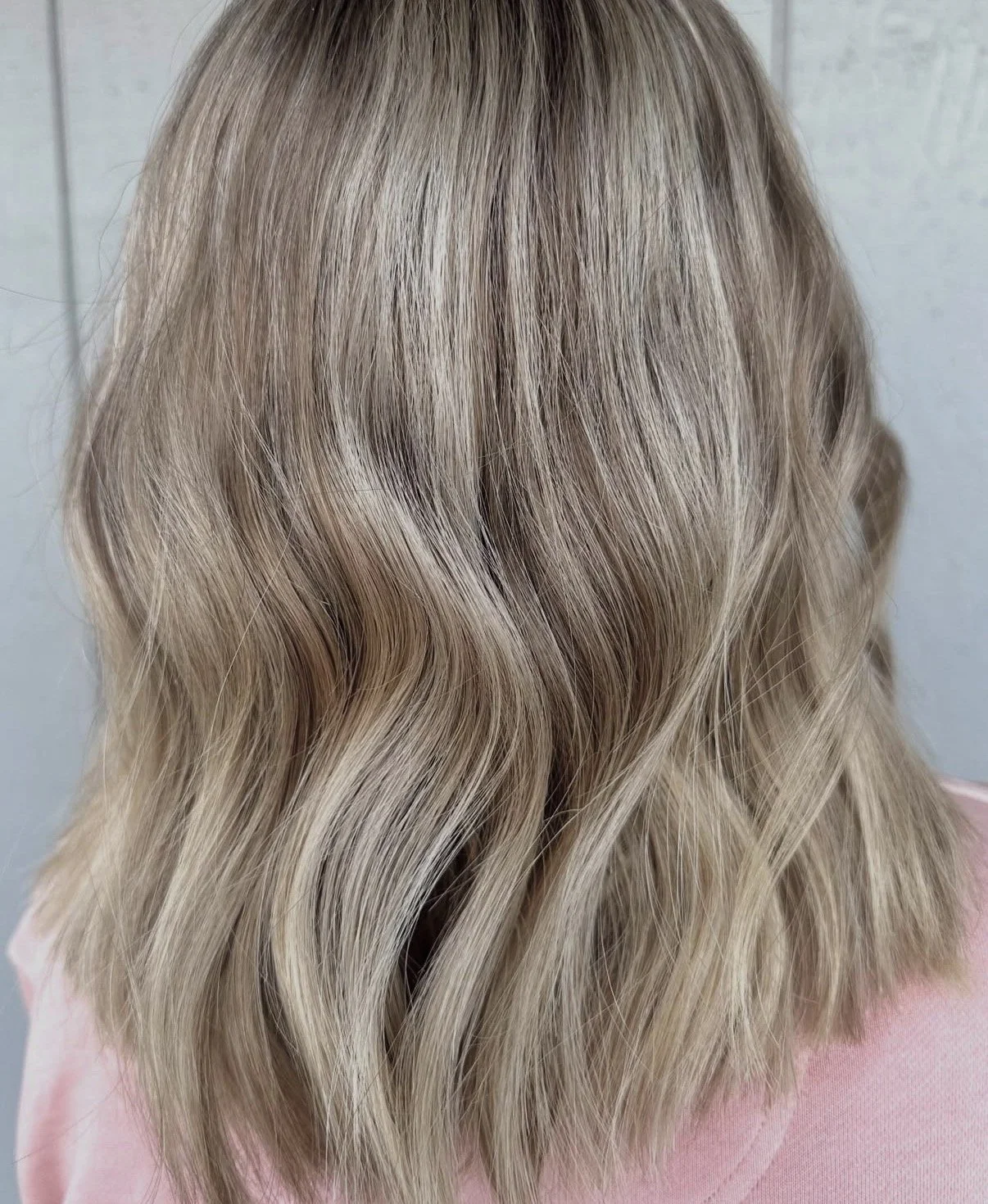 Back of a person's head with shoulder-length wavy blonde hair, wearing a pink shirt.