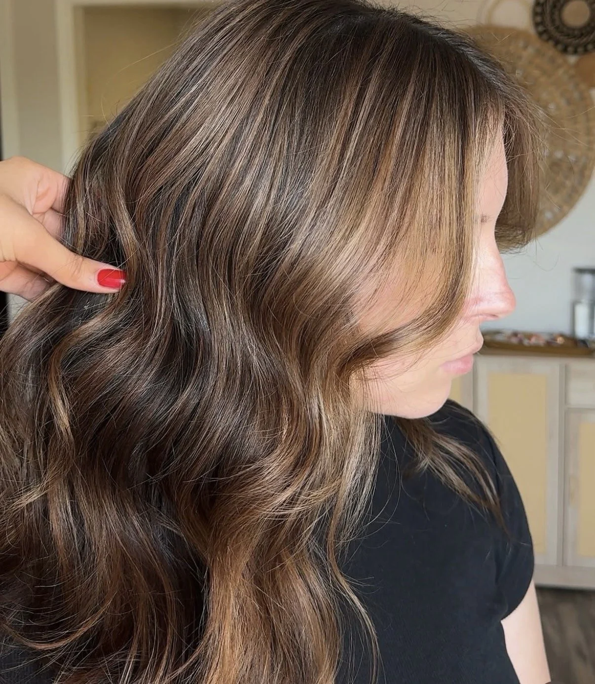 Side view of a woman with wavy, brown hair and blonde highlights, sitting in a room with a light-colored wall and decorative round wall hangings.