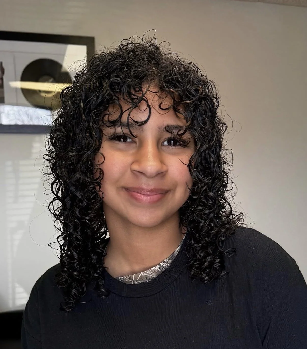 A woman with curly black hair smiling at the camera, wearing a black shirt in an indoor setting.