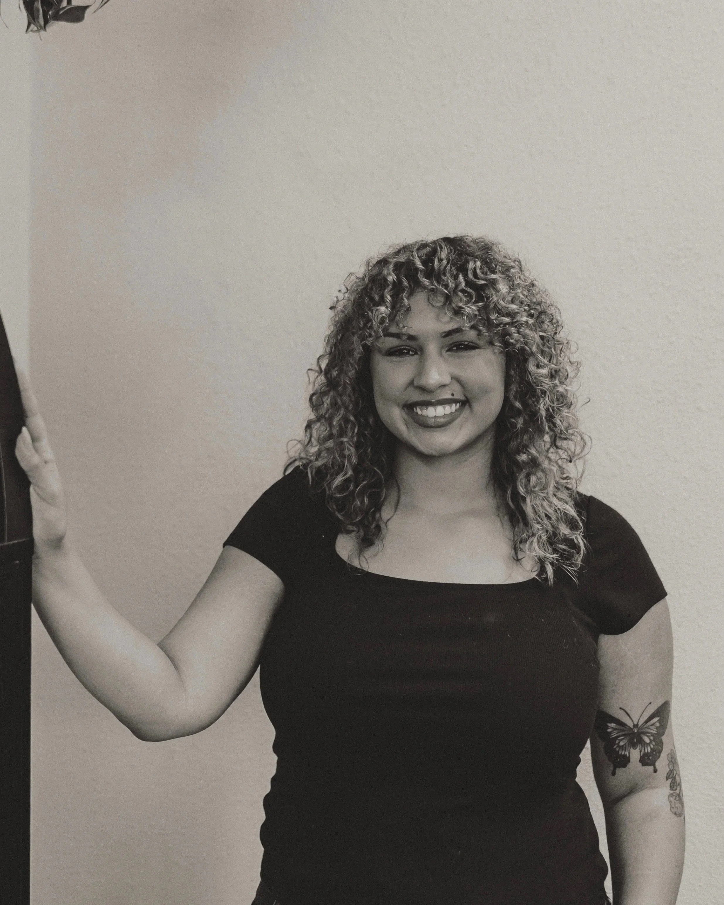 Smiling woman with curly hair, tattoos on her arm, wearing a black shirt, standing against a textured wall.