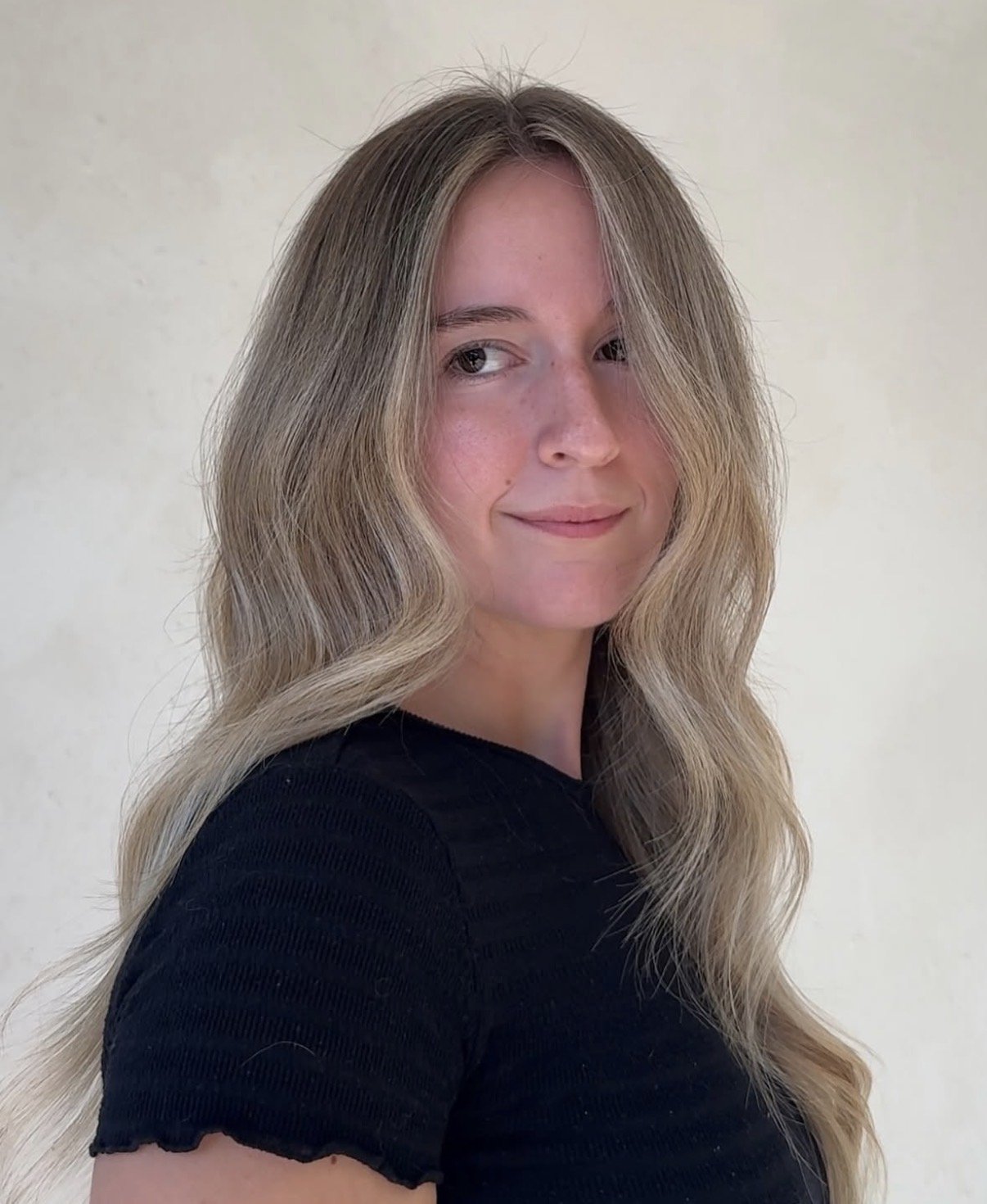 A young woman with long, wavy blonde hair, wearing a black top, poses against a plain background.