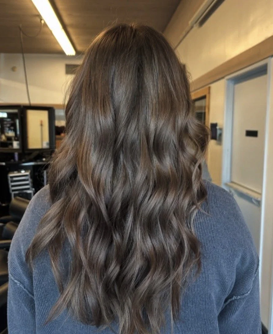Back view of a person with long, wavy, brown hair inside a salon.