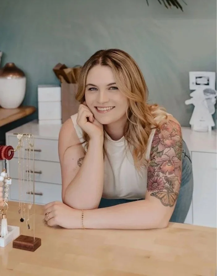 A smiling woman with blonde hair and a tattoo sleeve on her right arm, sitting at a wooden front desk  table with jewelry displayed on a stand, in the salon with white cabinets and decorative items in the background.
