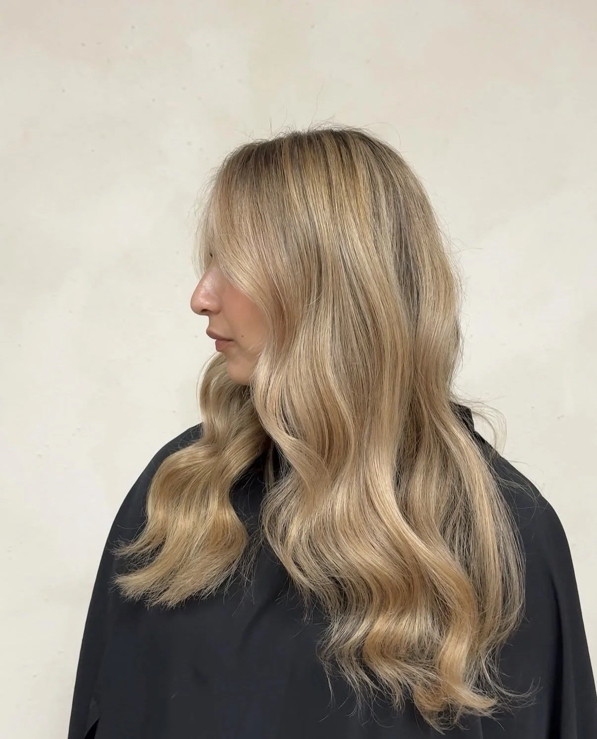 A woman with long, wavy blonde hair looking to the side, wearing a black top, against a plain light background.