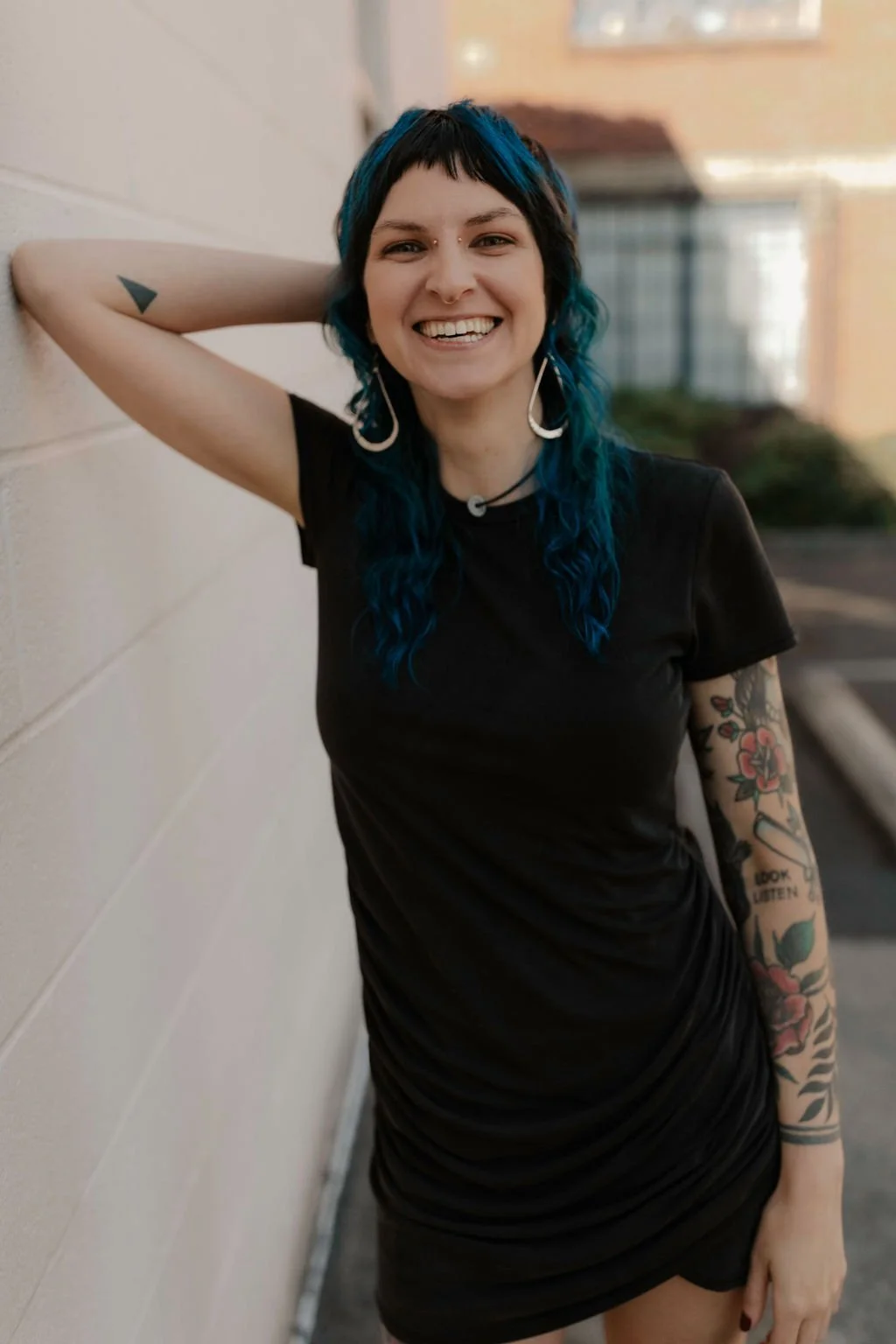 A smiling woman with black and blue hair, tattoos on her right arm, wearing a black dress, standing outdoors against a beige brick wall.