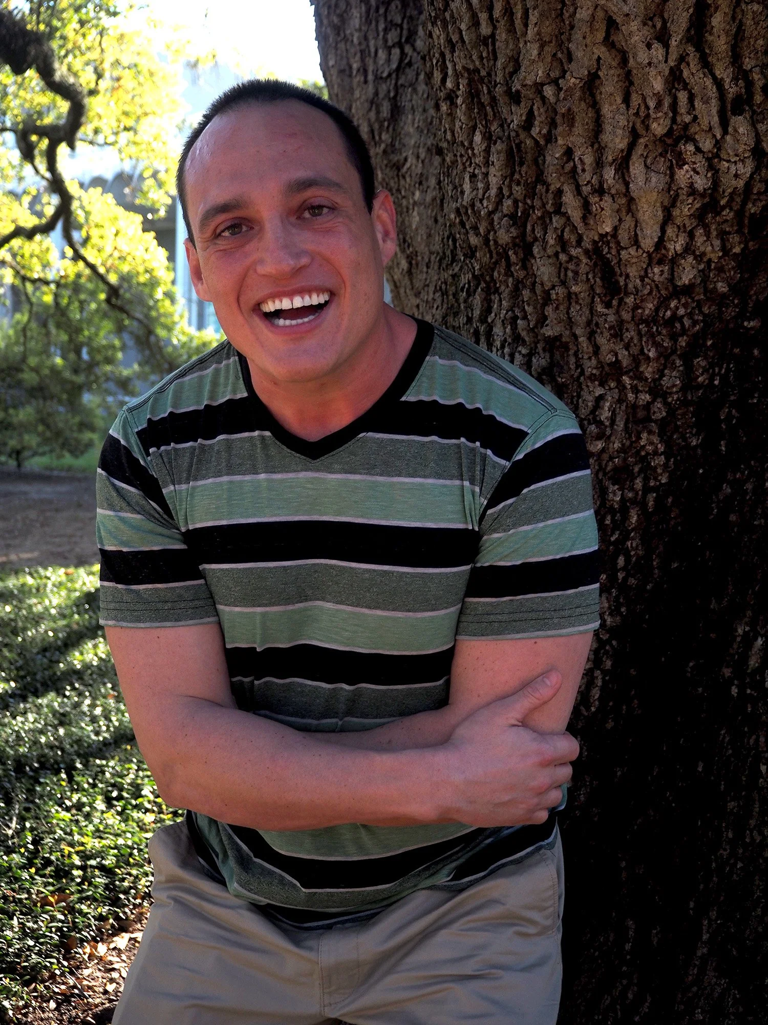 Cay smiles, leaning against a tree in a striped shirt.