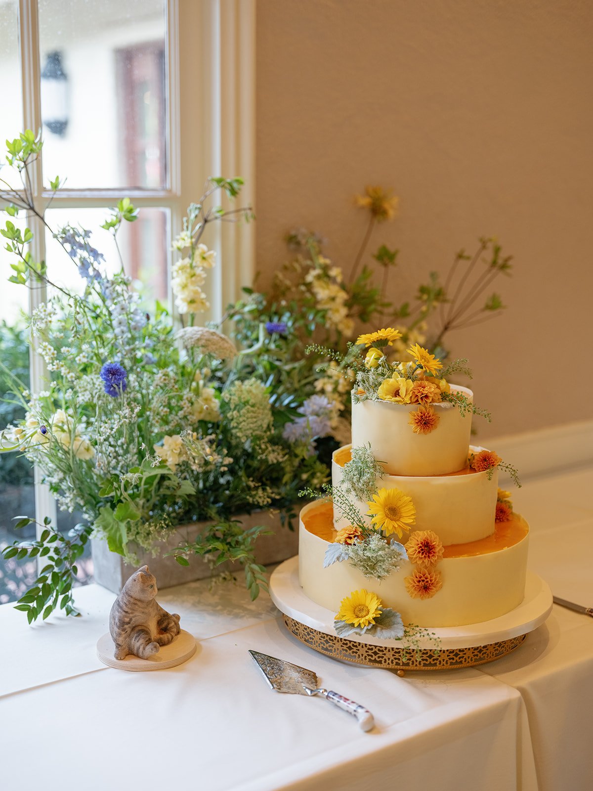 Three-tier yellow wedding cake decorated with sunflowers and orange flowers beside a floral arrangement and cat figurine