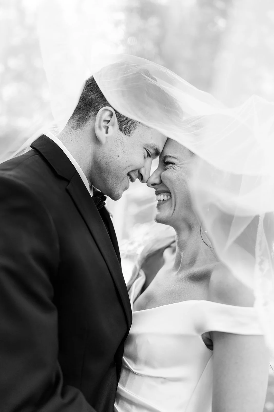 Black-and-white wedding portrait of a smiling bride and groom touching foreheads beneath her veil