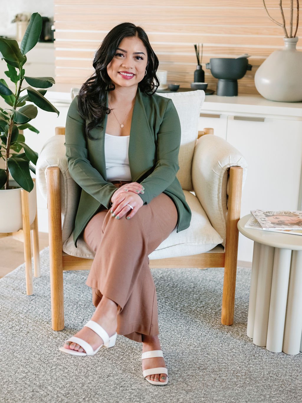 Smiling woman in a green blazer and tan pants sitting in a modern armchair beside a houseplant