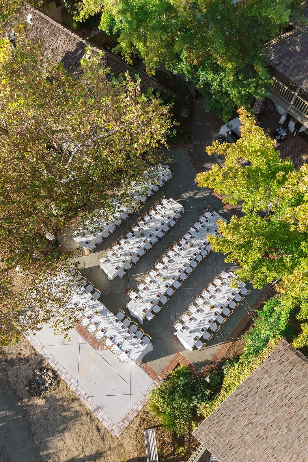 Aerial view of an outdoor restaurant patio with long tables set for a large event, surrounded by trees and rustic buildings.