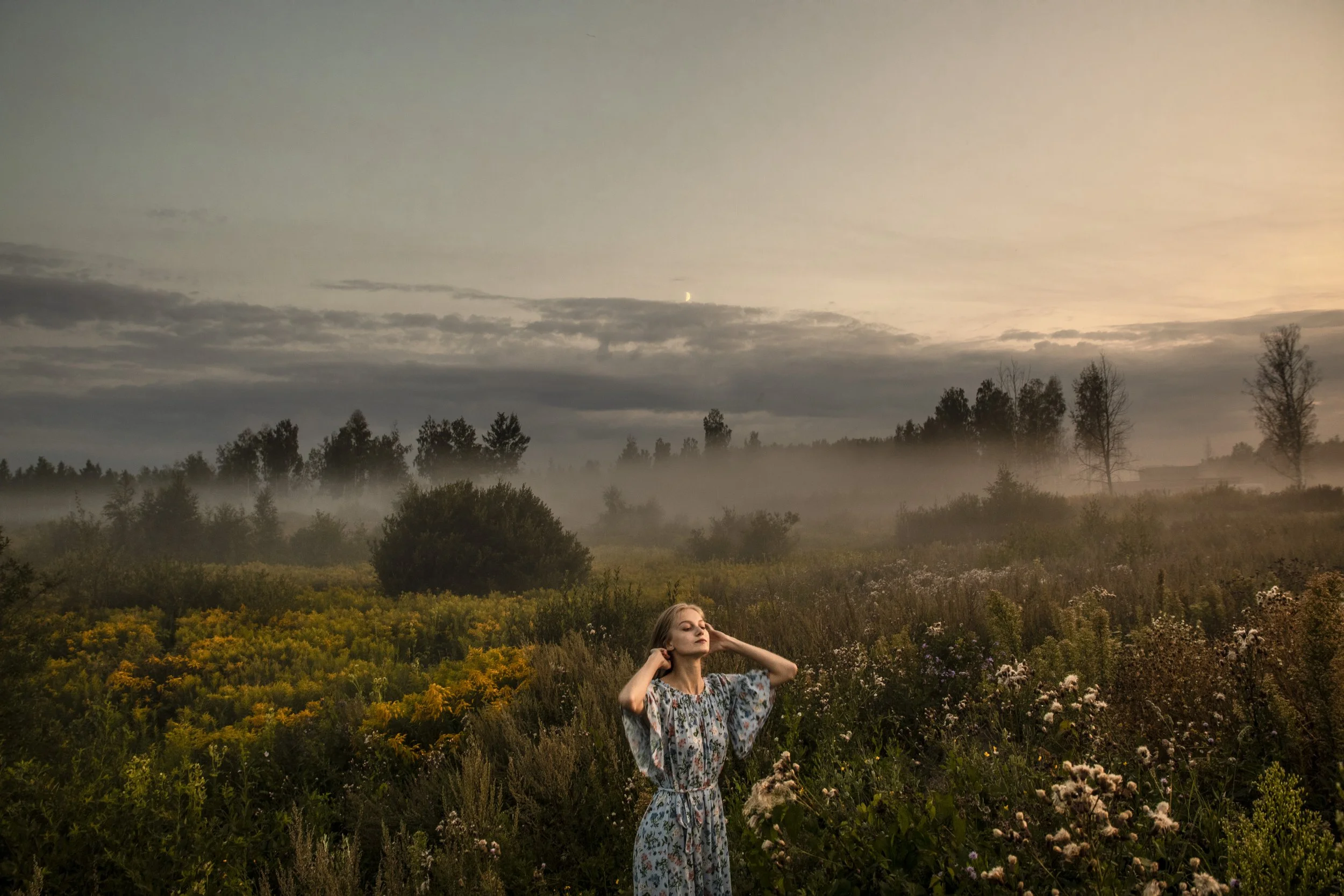 A woman standing in a field with tall grass and flowers, with her eyes closed and hands on her head, under a cloudy sky during sunset.