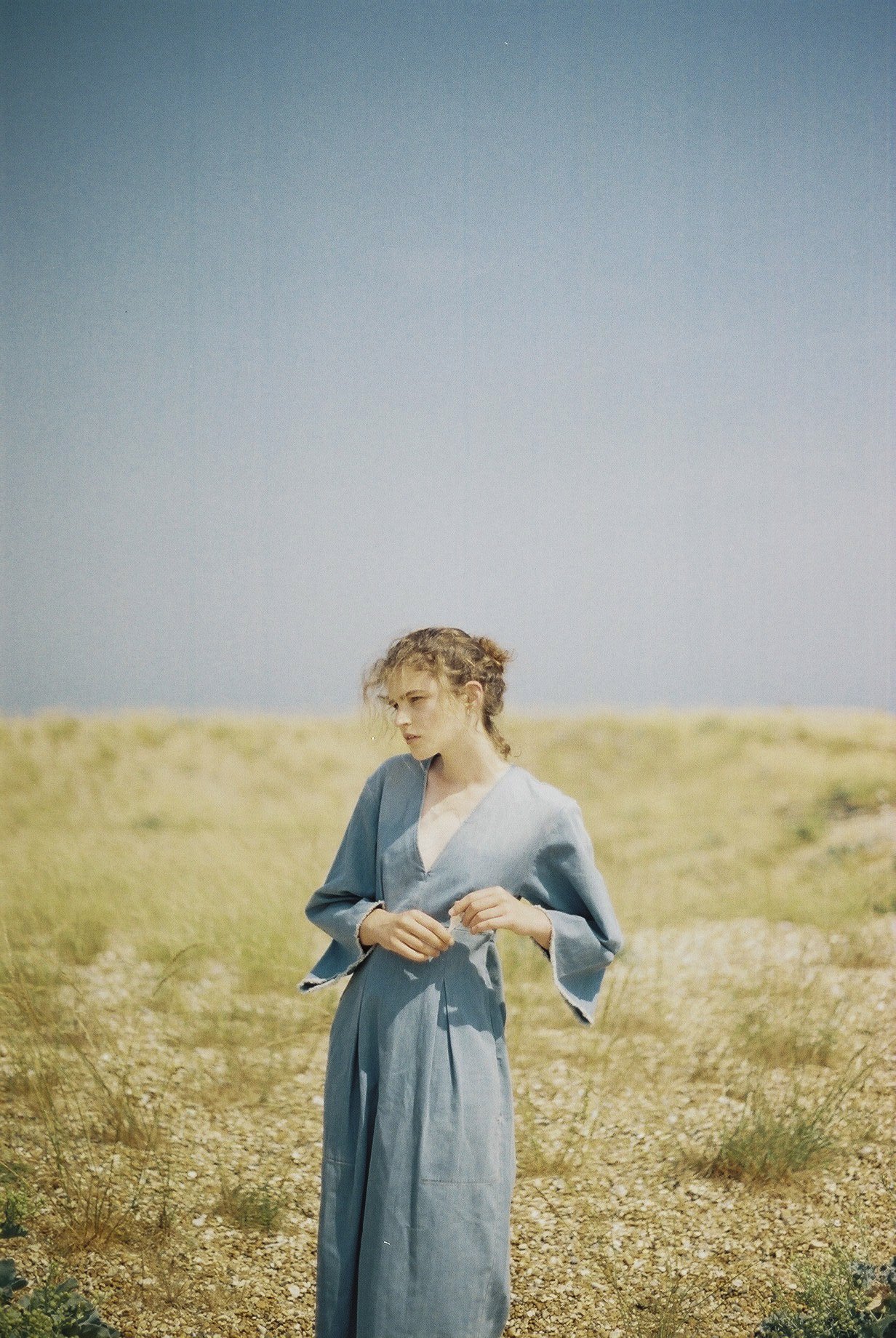 A woman with curly hair stands outdoors in a grassy, rocky area under a clear blue sky, wearing a long blue dress.