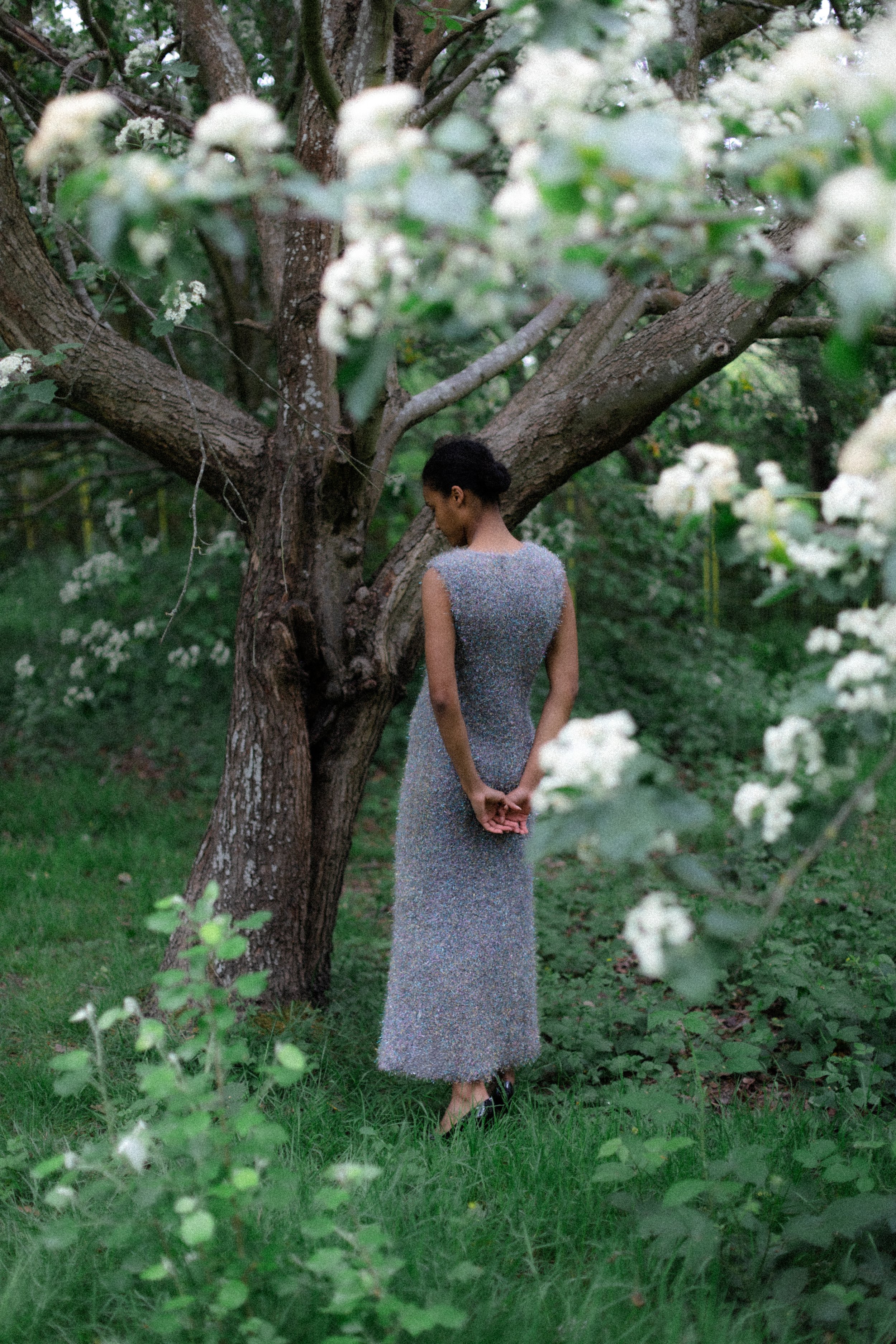 A woman in a sparkly silver dress standing outdoors near a large tree with white flowers and green foliage.