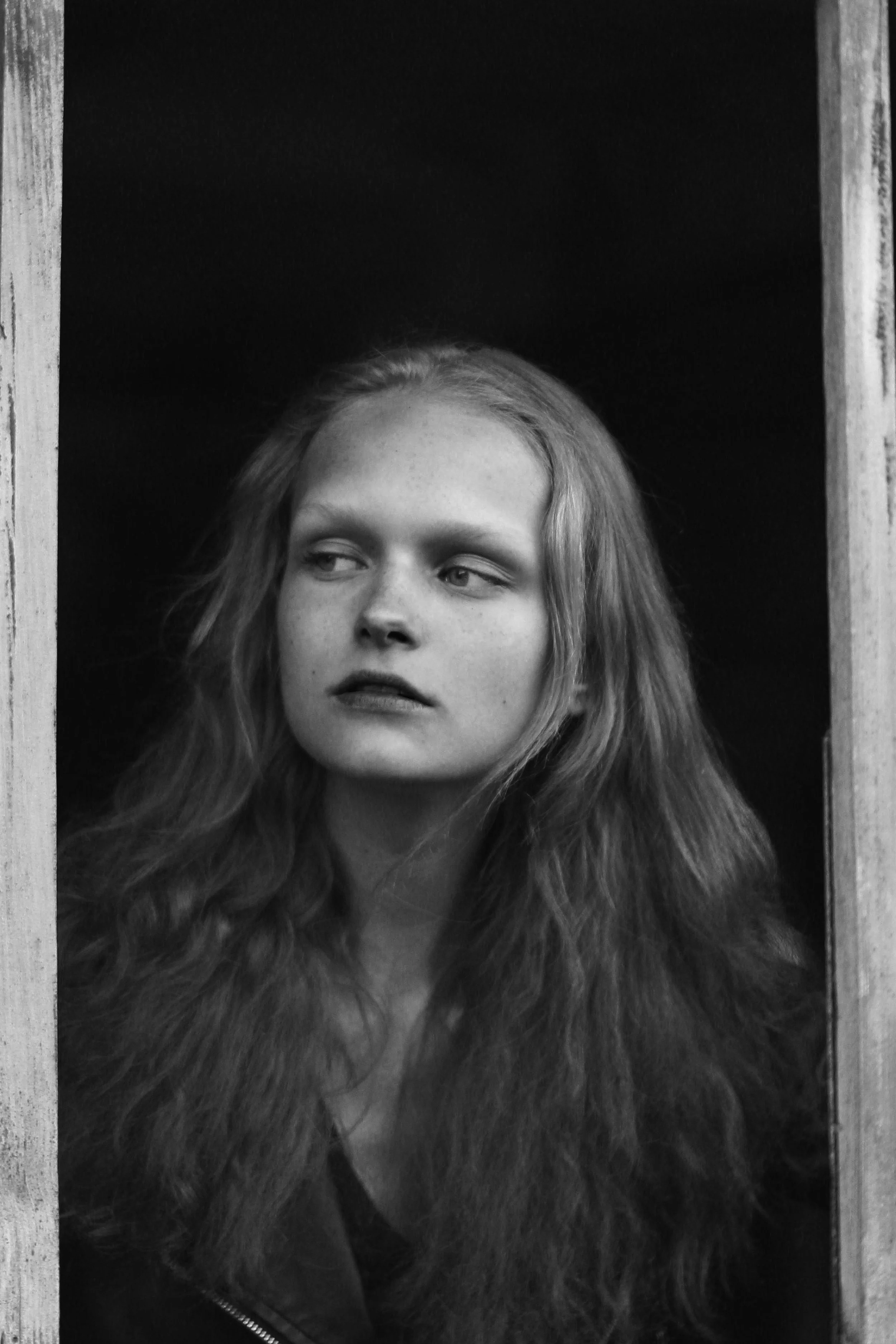 Black and white portrait of a young woman with long wavy hair looking to her right, framed by wooden borders, wearing a jacket with a zipper.