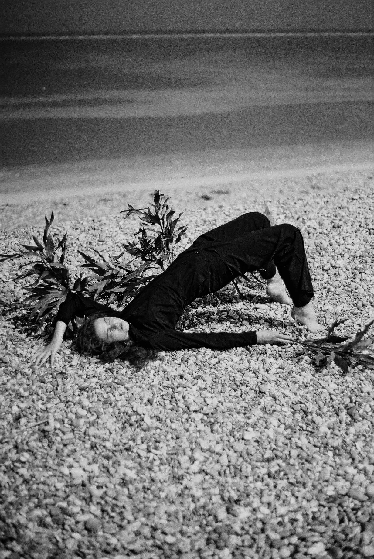 A woman lying on a pebble beach with her eyes closed, wearing dark clothing, with a branch or plant near her head.